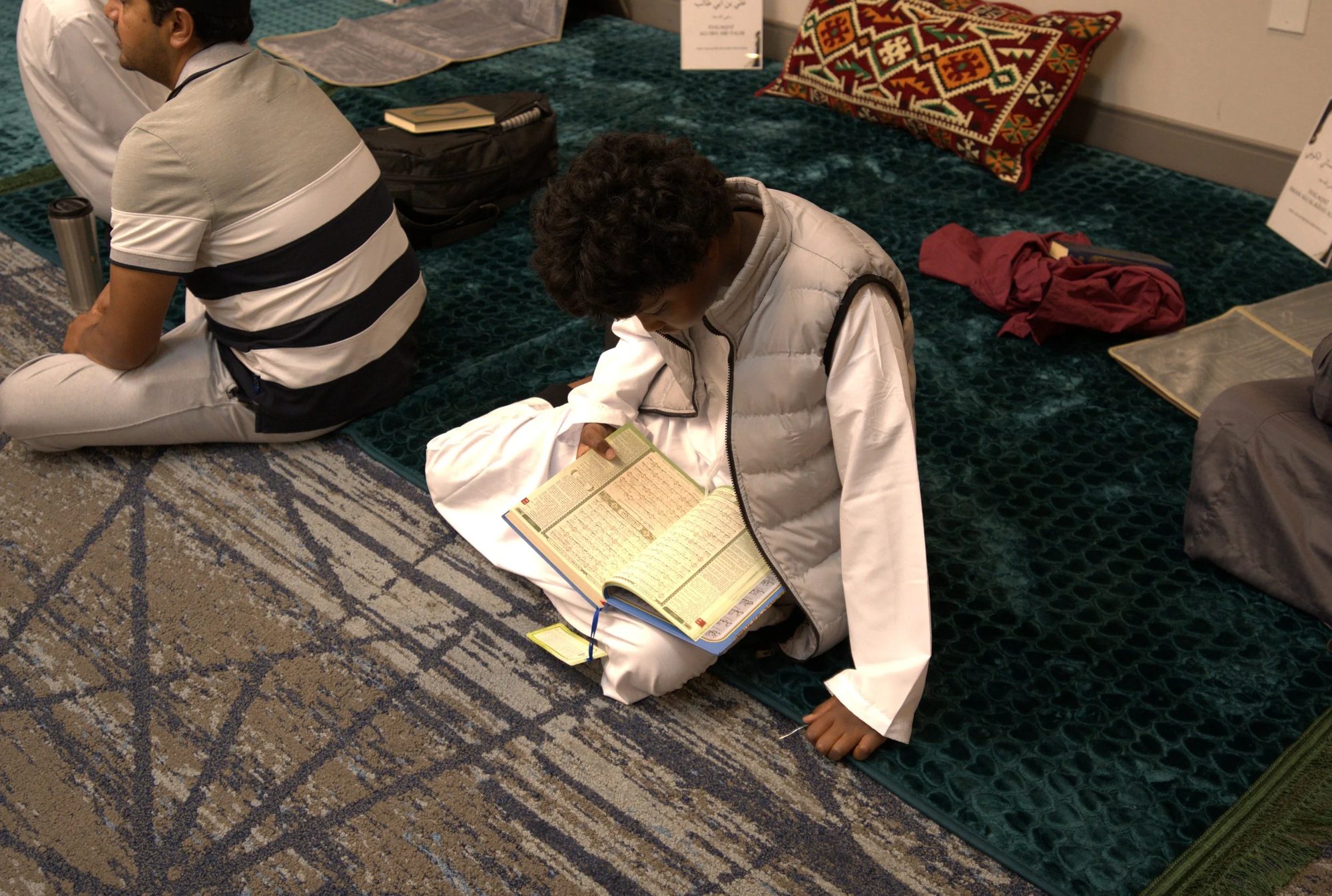 A young student reads from an open mushaf while seated on the prayer hall carpet