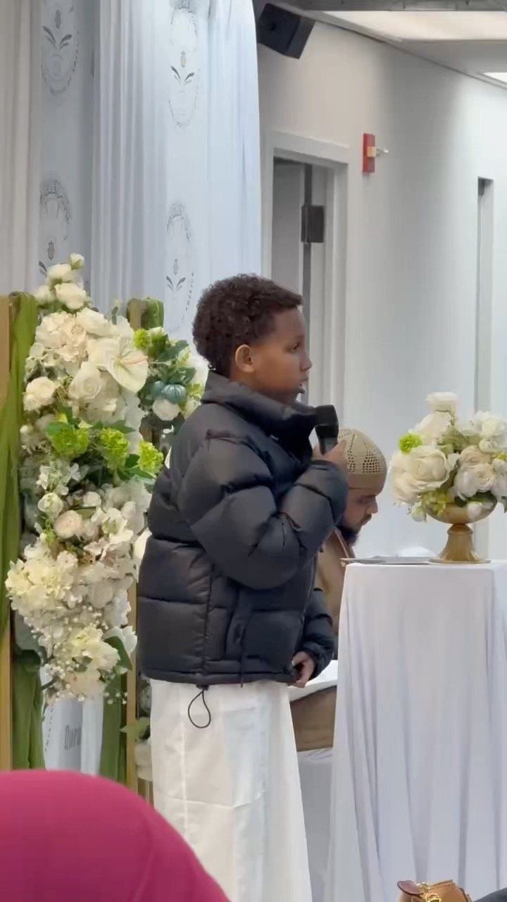 A boy in a black jacket speaks into a microphone beside white flower arrangements