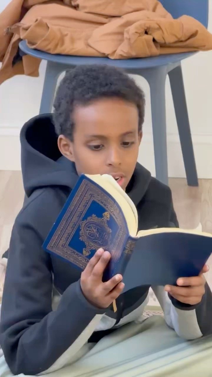 A boy reads aloud from a blue mushaf held close to his face