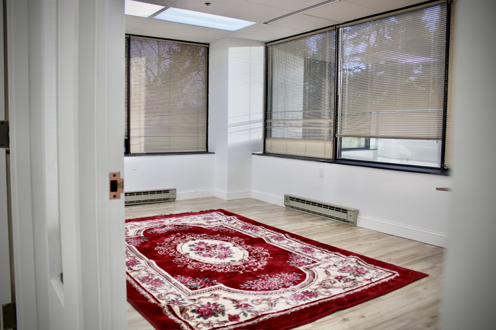 A red patterned prayer rug on the wooden floor of a bright room with windows