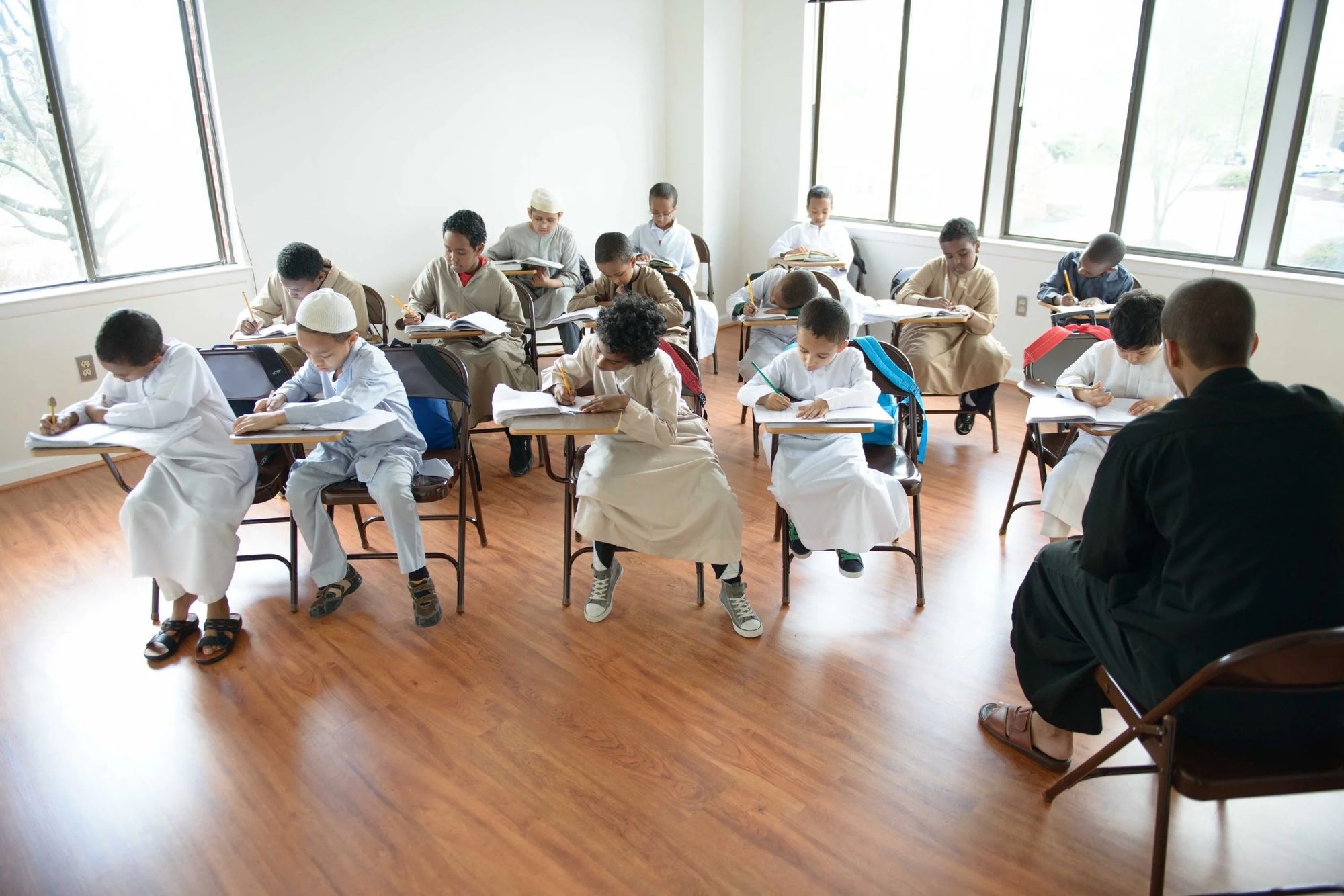 Boys in white thobes seated at desks with open mushafs in a sunlit classroom at the institute