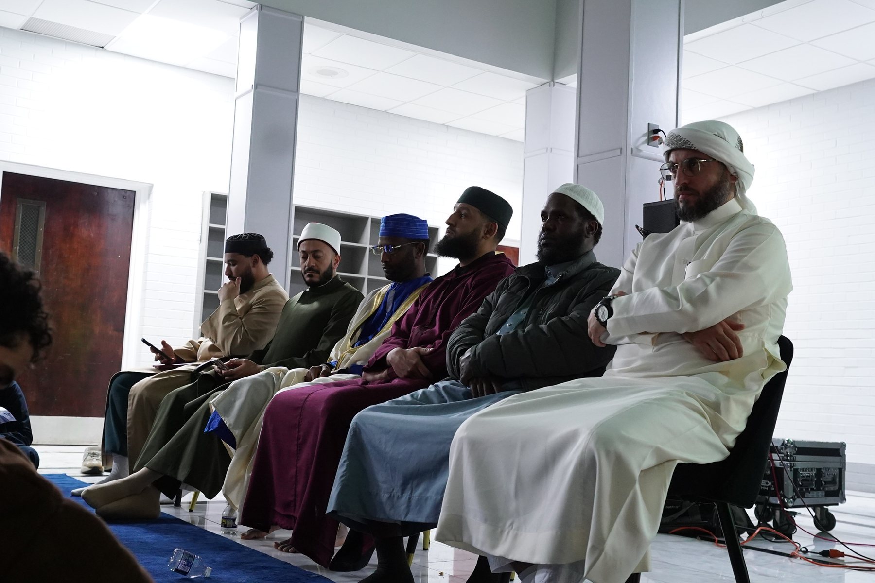 Six mashayikh in traditional dress seated in a row of chairs in a bright hall