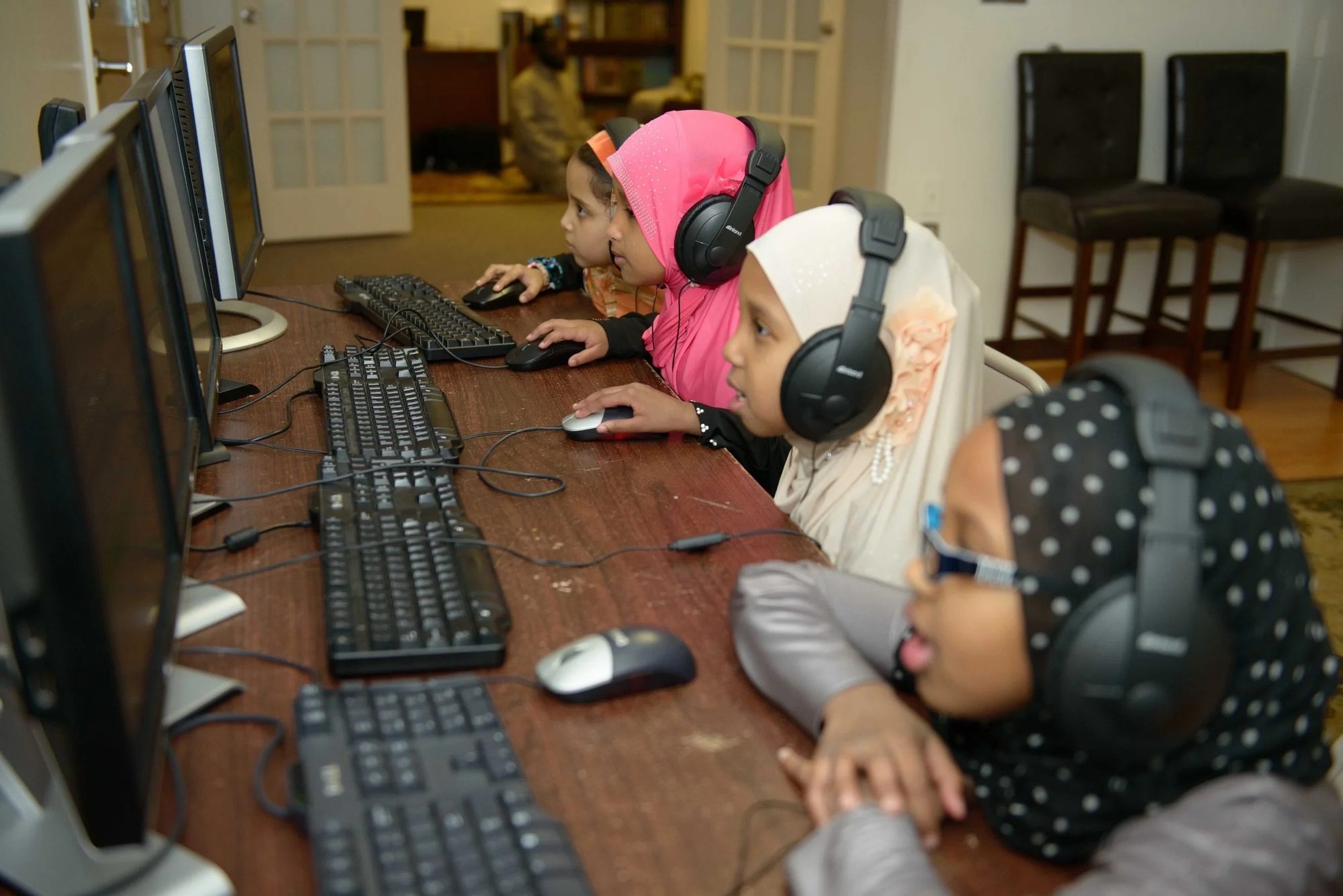Girls in hijab wearing headphones at computer stations during a Qur'an lesson