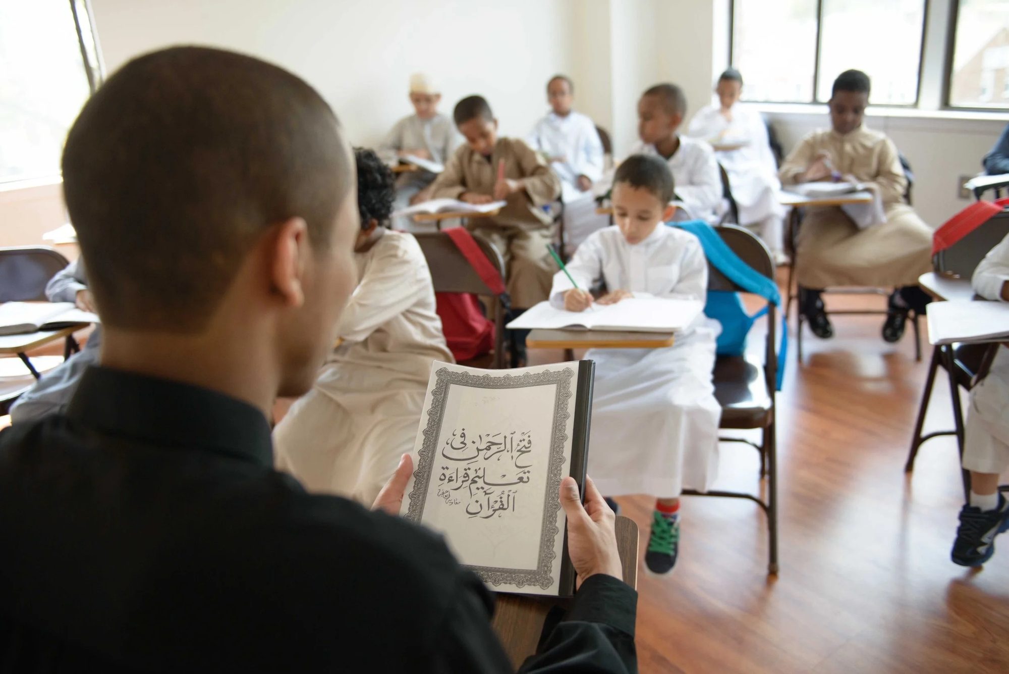 A teacher holds up a framed Qur'anic calligraphy while boys at desks look on